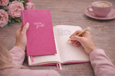 Donna che scrive su un taccuino A5 rosa “Think Pink!” su una scrivania, scena lifestyle elegante e naturale per lavoro e creatività